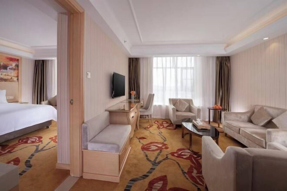 Quarto de hotel com cama, sofá, poltrona, mesa de centro e televisão.