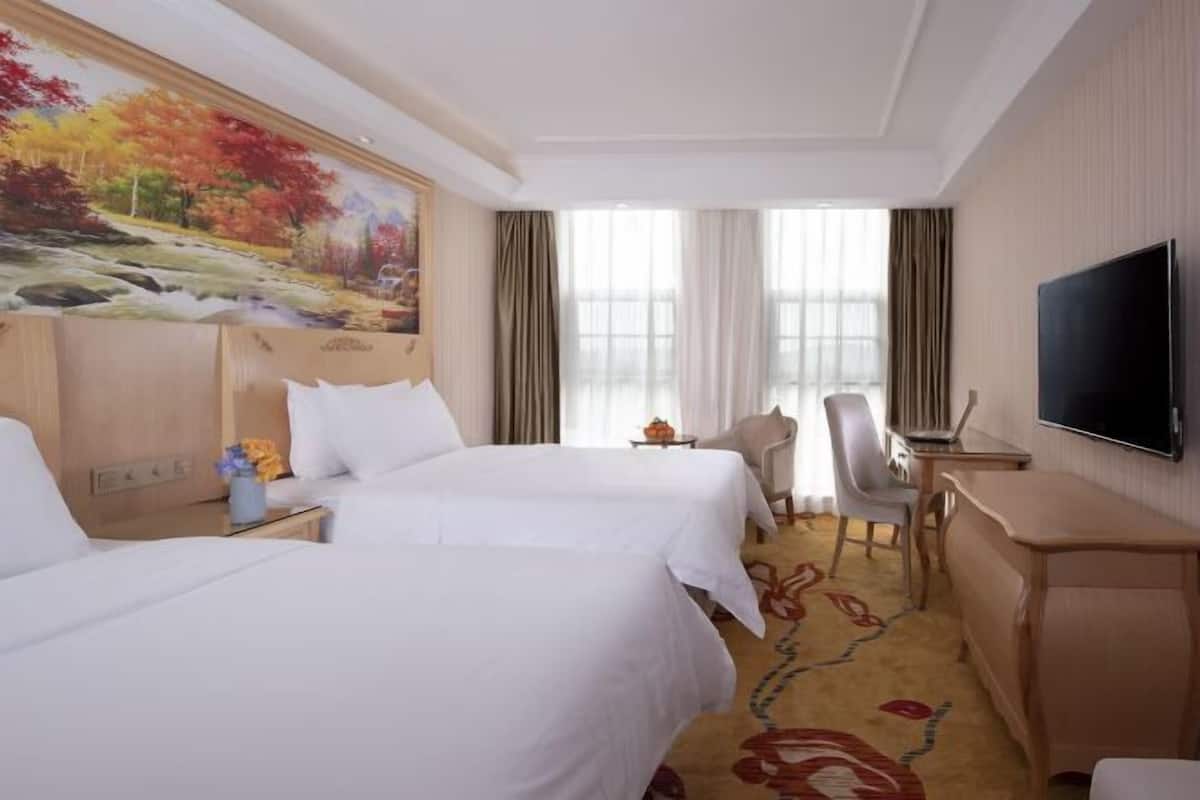 Quarto de hotel com duas camas, uma escrivaninha, uma cadeira, uma televisão e um quadro na parede.