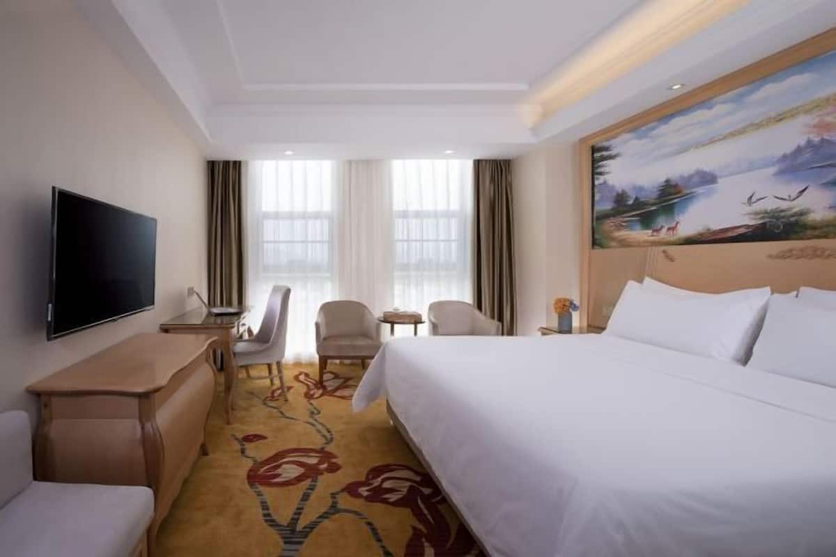 Quarto de hotel com cama, televisão, escrivaninha, cadeira, mesinha e um quadro na parede.
