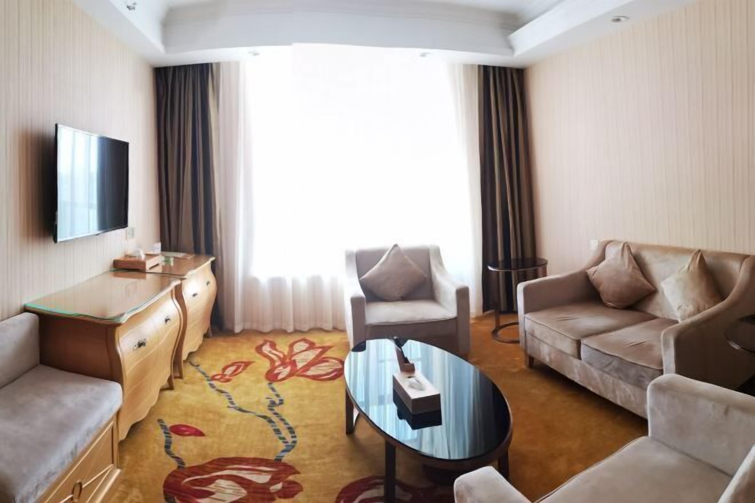 A hotel room with a flat-screen TV, a sofa, a chair, a coffee table, a desk, and a large window with curtains.