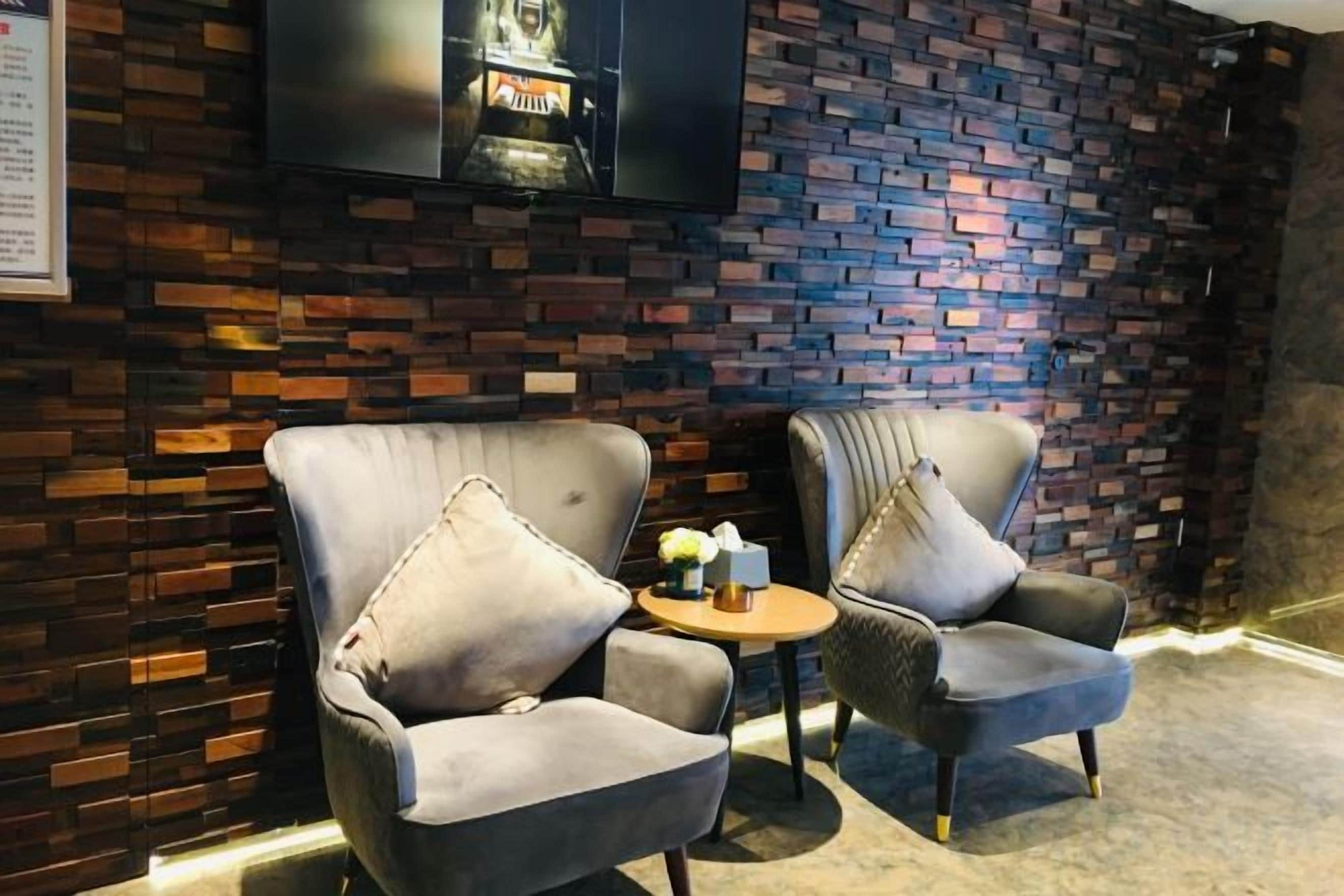 Two armchairs with cushions, a small round table with a vase and a book, and a flat-screen TV mounted on a brick wall.