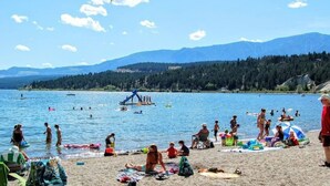 A beach with people sunbathing and swimming, a dock with boats, and mountains in the background. - Blue Heron Pointe | Lakeview | Pool | Hot Tub |Gym (Invermere)