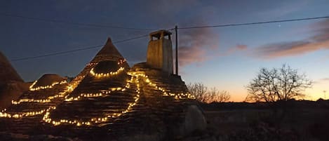 A stone structure with a conical roof and a chimney, illuminated by string lights.