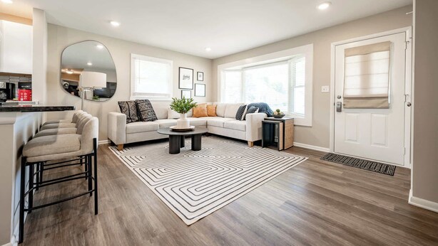 A modern living room with a sectional sofa, a coffee table, and a large mirror. There is a kitchen area visible to the left. - Speedway Luxe | Hot Tub | Shuffle | Steps to Track (Indianapolis)