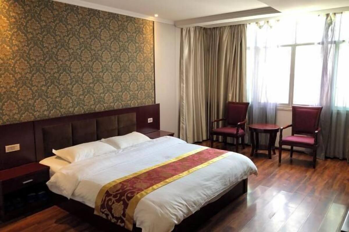 Een hotelkamer met een groot bed, een houten hoofdbord, behang met een patroon, een raam met gordijnen en twee stoelen.