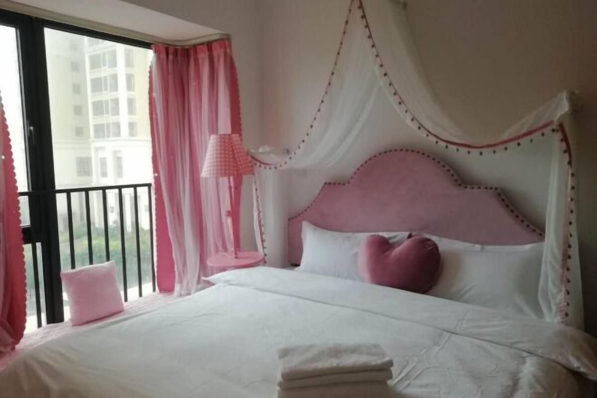 Une chambre à coucher avec un lit, des rideaux roses et une vue sur les bâtiments environnants.