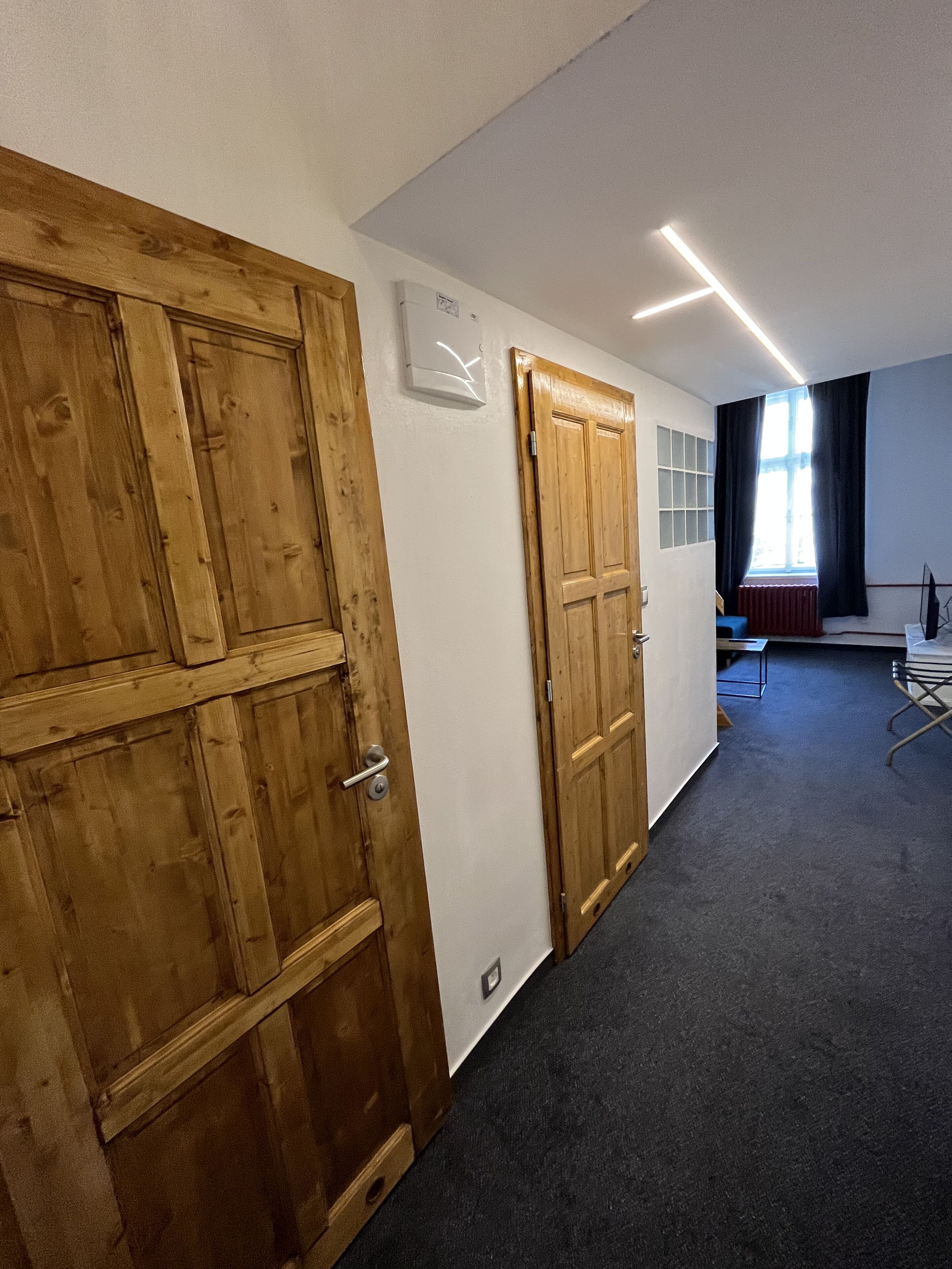 A hallway with wooden doors, a white wall with a light switch, a radiator, and a window with curtains.
