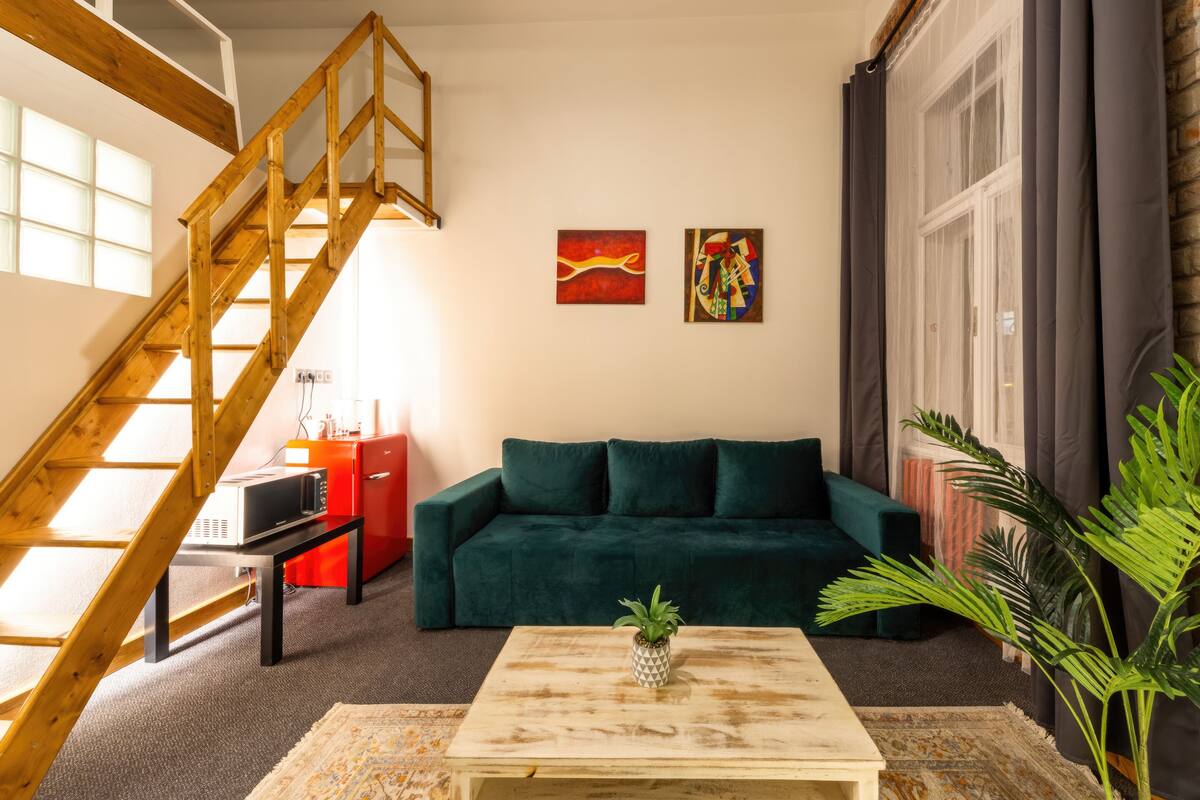 Ein Wohnzimmer mit einem grünen Sofa, einer Holztreppe, einem Couchtisch mit Pflanze und zwei Kunstwerken an der Wand.