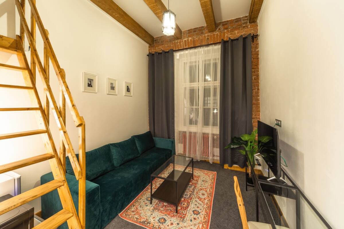 Ein Wohnzimmer mit einer Holztreppe, einem grünen Sofa, einem Glastisch und einem Fernseher auf einem Schrank.