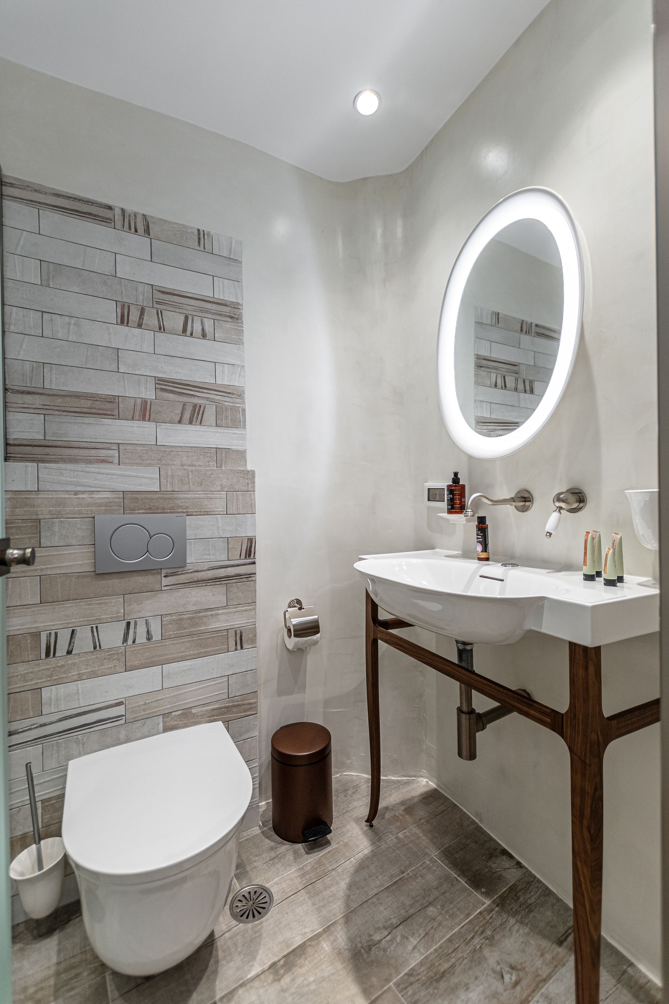 Un baño moderno con un inodoro blanco, un tocador de madera con espejo y una pared revestida de azulejos.