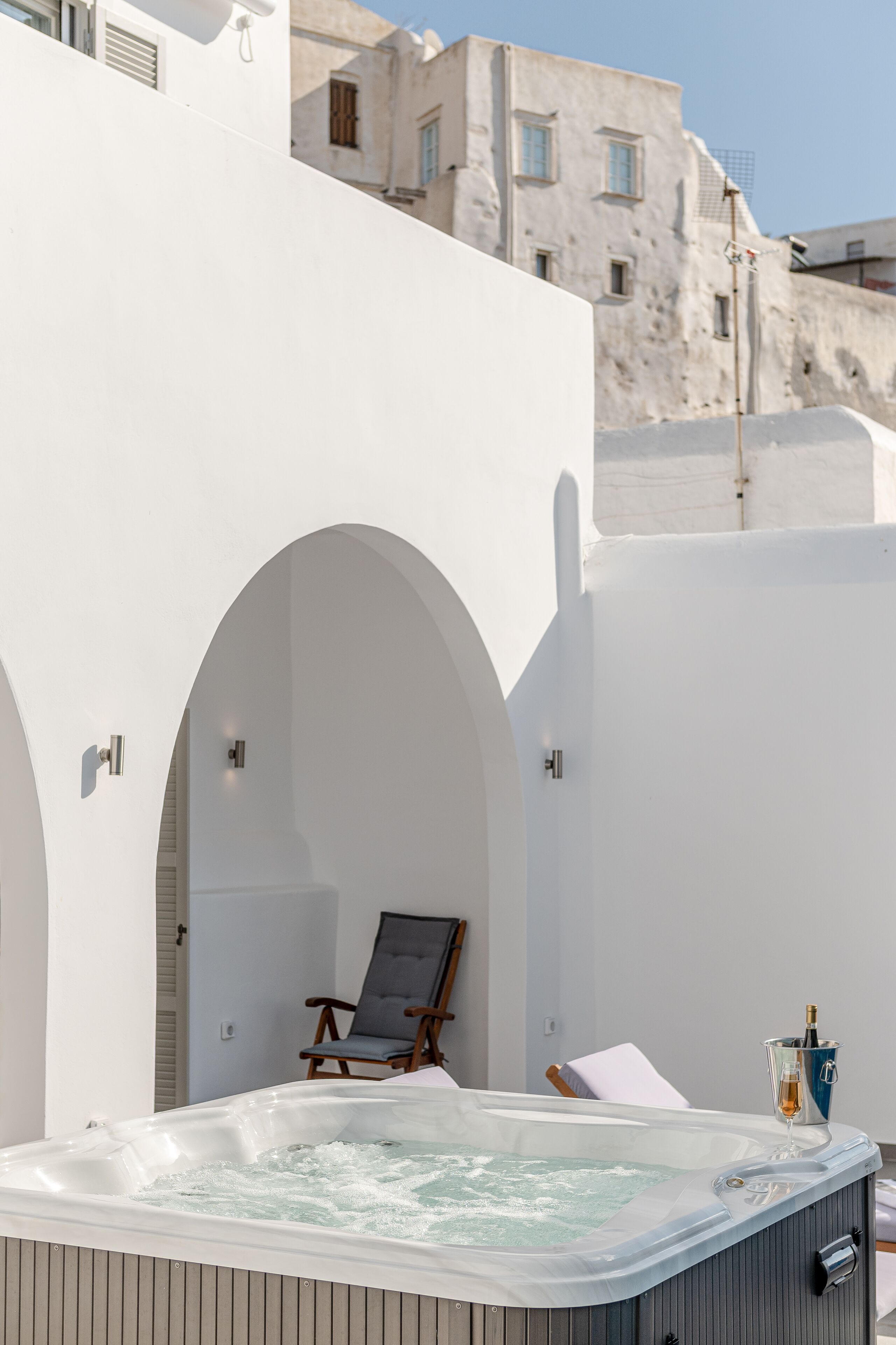 A hot tub with a wooden chair and a bottle of champagne, set against a white building with arched doorways.