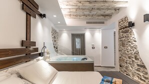 A modern hotel room with a wooden headboard, a large bed, a built-in hot tub, stone wall accents, and a view of the hallway. - Mariet Naxos Elegant Suites (Naxos)