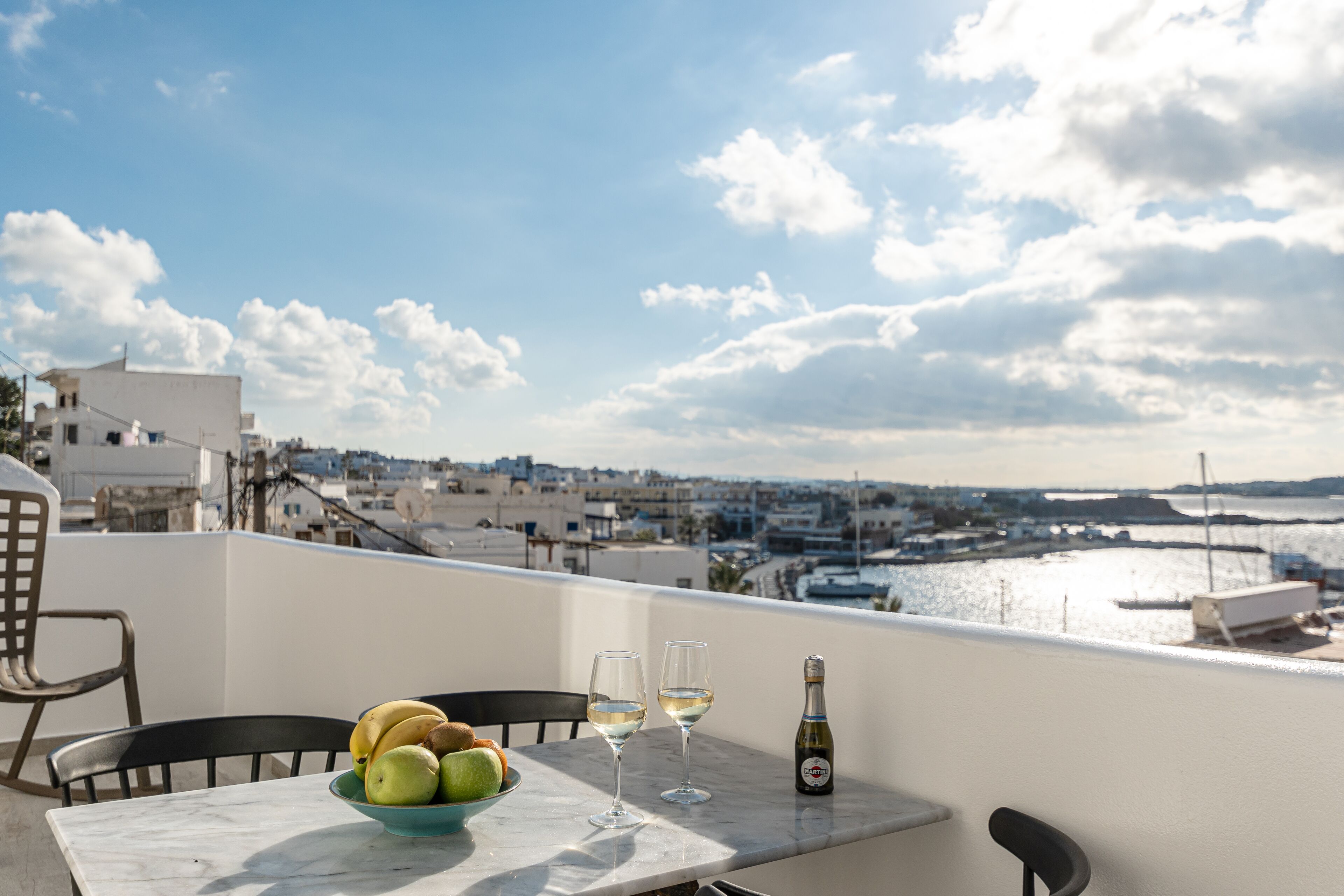 A terrace with a table, chairs, a bowl of fruit, two glasses of white wine, and a bottle of champagne.