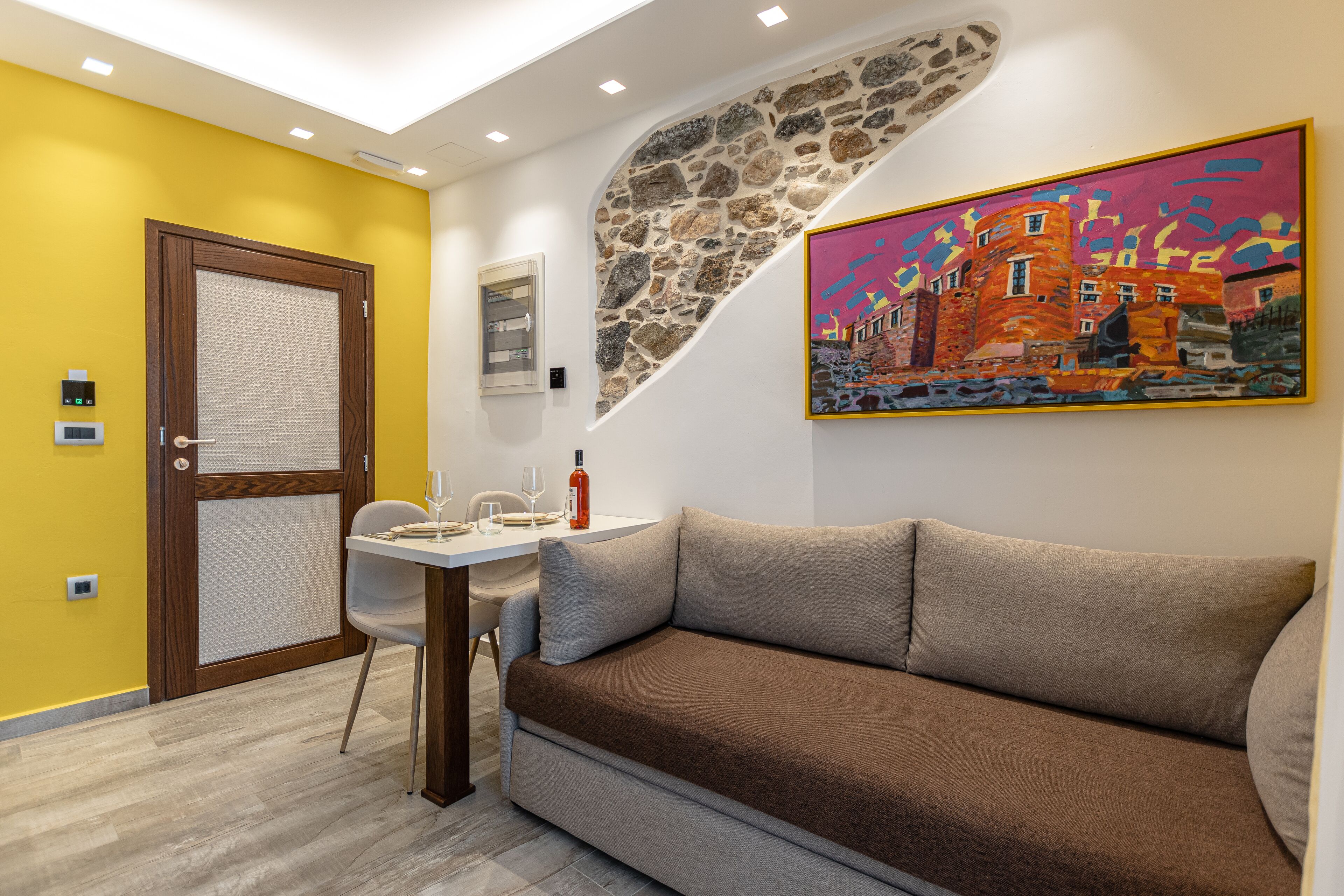 A room with a brown sofa, a dining table with chairs, a painting on the wall, and a stone accent wall.