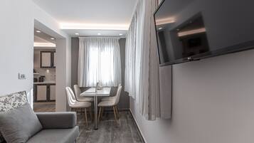 Uma sala de estar moderna com sofá, uma mesa de jantar com cadeiras, uma TV de tela plana na parede e uma área de cozinha ao fundo.