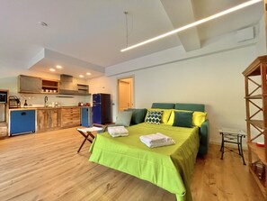 Una habitación pequeña con cama, una kitchenette, piso de madera y una lámpara en el techo.