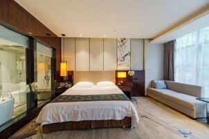Habitación de hotel con una cama grande, un sofá, un escritorio y un baño con cabina de ducha de vidrio.