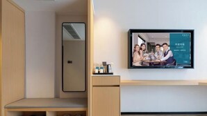 Hotellihuone, jossa on seinälle asennettu televisio, puinen penkki, jossa on kenkiä, ja pieni pöytä, jossa on hygieniatarvikkeita.