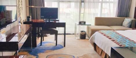 Una habitación de hotel con una cama, un escritorio con una silla, un televisor y un ventanal con cortinas.