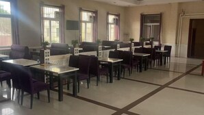 Comedor de hotel con varias mesas y sillas, ventanas amplias con cortinas y suelo de baldosas.