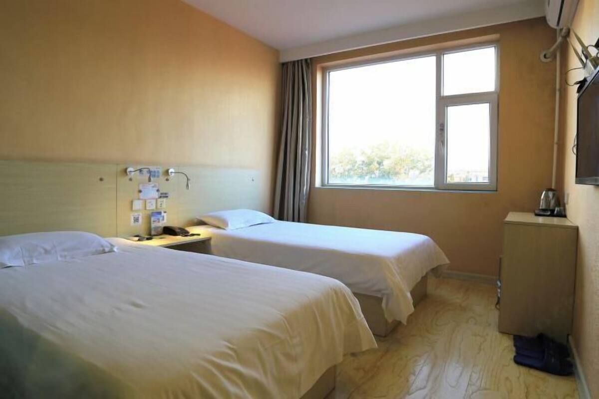 A hotel room with two beds, a bedside table with a lamp, a small cabinet, and a window with curtains.