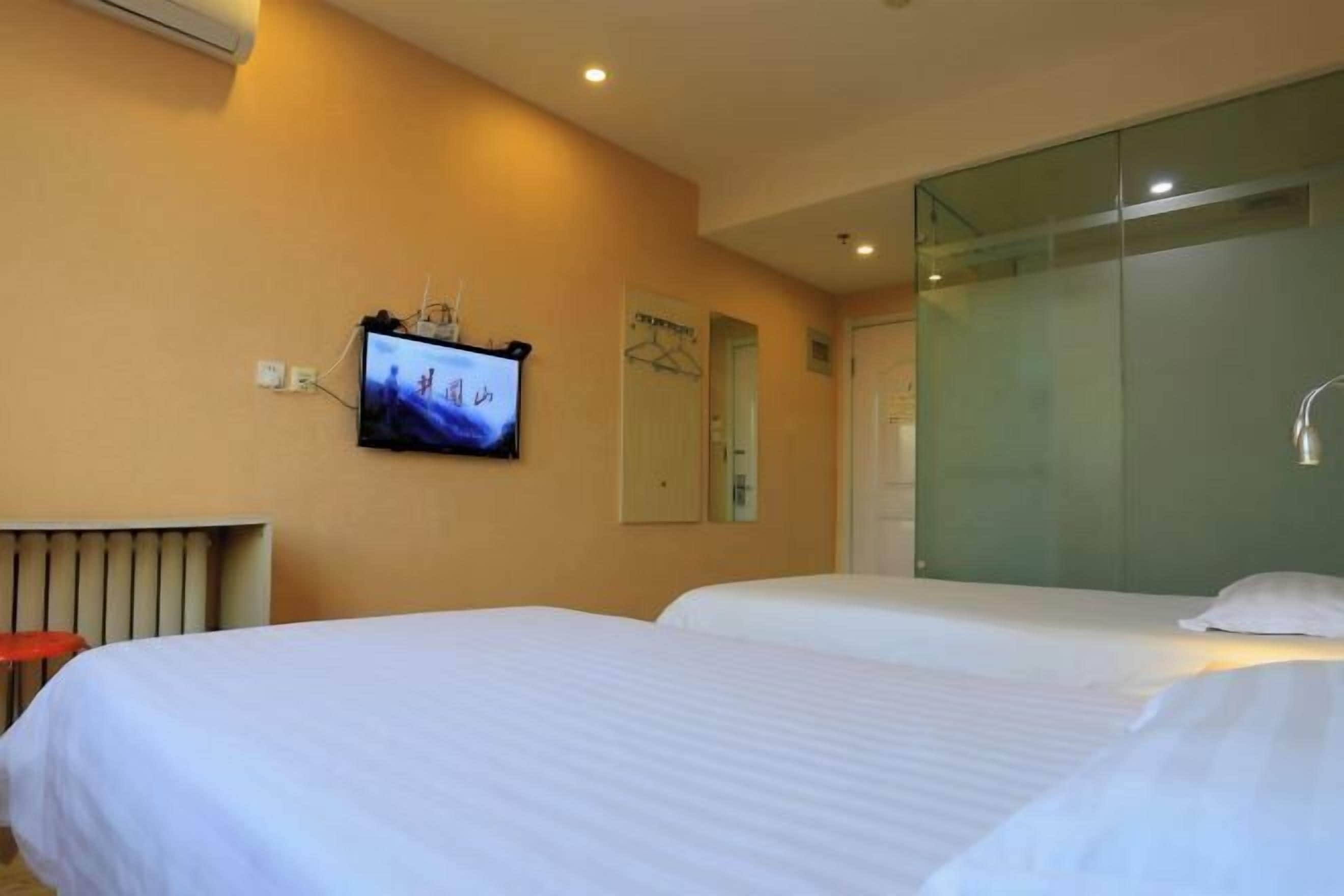 Ein Hotelzimmer mit zwei Betten, einem an der Wand befestigten Fernseher und einer Glastrennwand, die den Raum vom Badezimmer abtrennt.