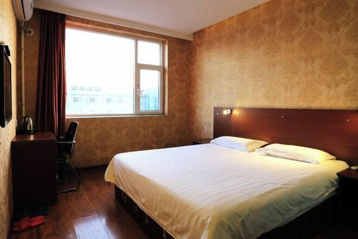 A hotel room with a bed, a desk, a chair, a window, and a wall-mounted air conditioning unit.
