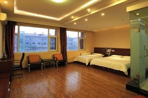 Habitación de hotel con dos camas, zona de estar con sillas, un ventanal con vistas a edificios y un panel de vidrio.
