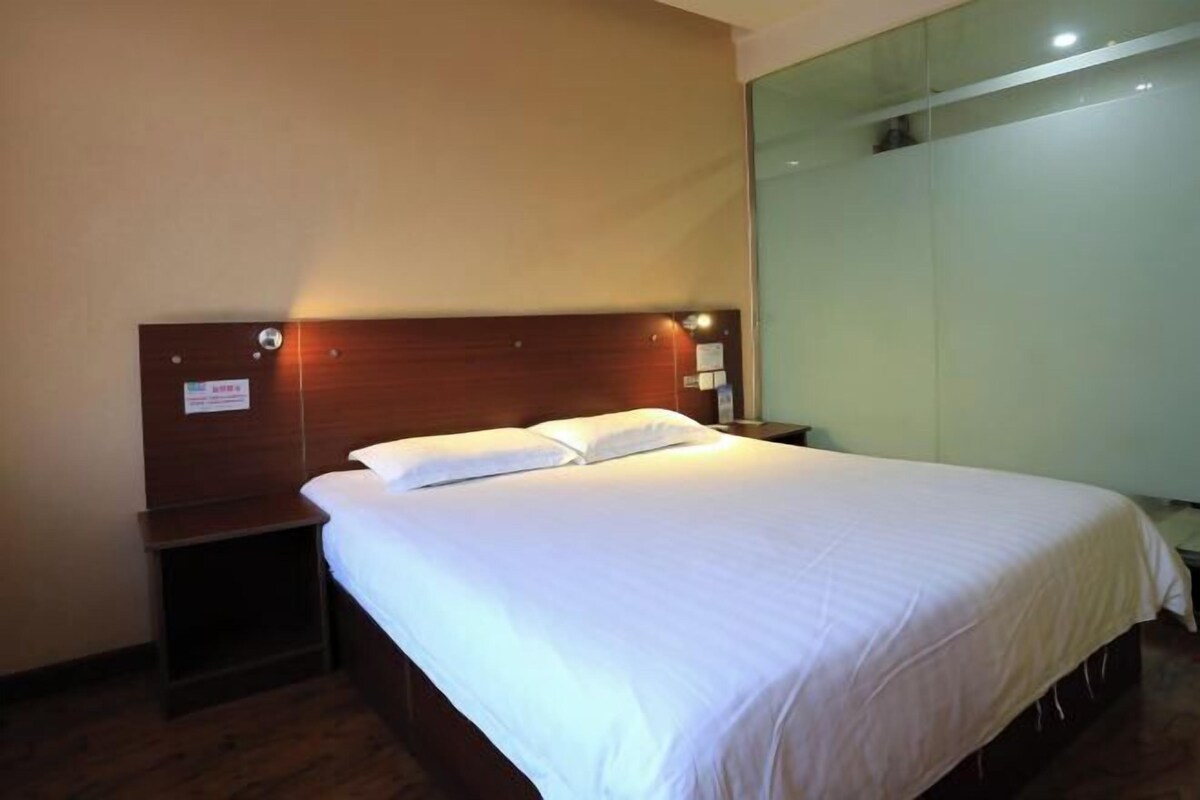 Habitación de hotel con cabecera de madera, una cama individual con ropa blanca, una mesita de noche y una lámpara de pared.
