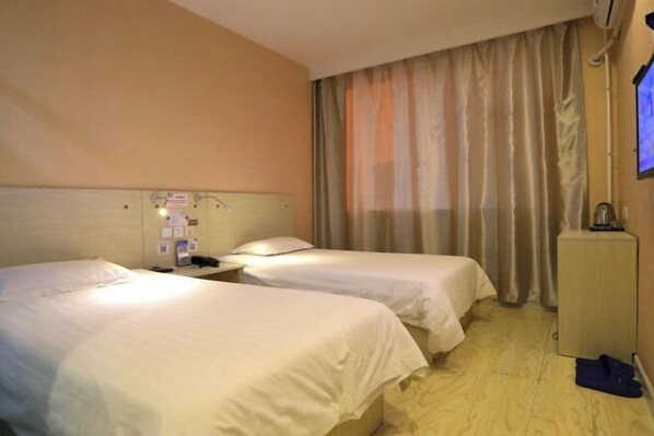 A hotel room with two single beds, a bedside table with a lamp, a small cabinet, and a TV mounted on the wall.