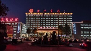 A hotel with a large, illuminated sign and multiple flags on top. There is a parking lot with several cars and a fountain in the center. - Chiping Hotel (Liaocheng)