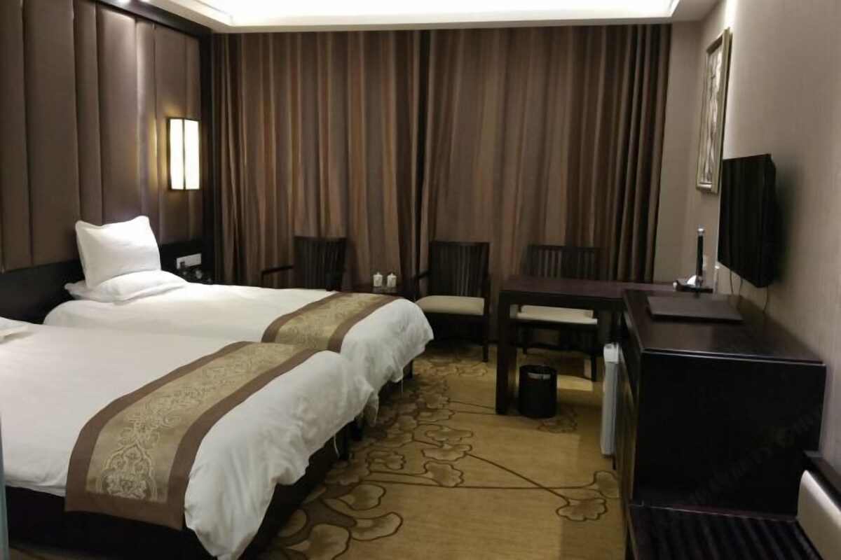 Habitación de hotel con dos camas, un escritorio, una silla, un televisor y un ventanal grande con cortinas.