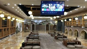A spacious lobby with a large TV screen displaying various content, wooden seating areas, and a tiled floor.