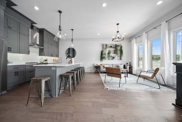 Une cuisine moderne avec des meubles gris, un îlot central doté de tabourets, un éclairage suspendu et un coin repas comprenant un canapé et des chaises.