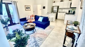 Ett modernt vardagsrum med en blå soffa, ett soffbord och en matplats med ett litet bord och stolar. Det finns också ett köksområde med vita skåp och ett kylskåp.