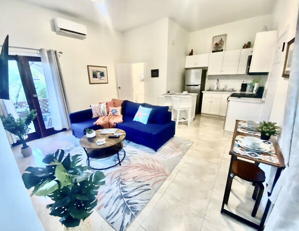 A modern living room with a blue sofa, a coffee table, and a dining area with a small table and chairs. There is a kitchen area with white cabinets and a refrigerator.