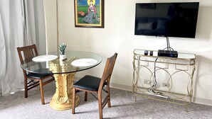 A dining area with a glass table, wooden chairs, a decorative pedestal, a chandelier, a framed painting, and a television on a gold stand.