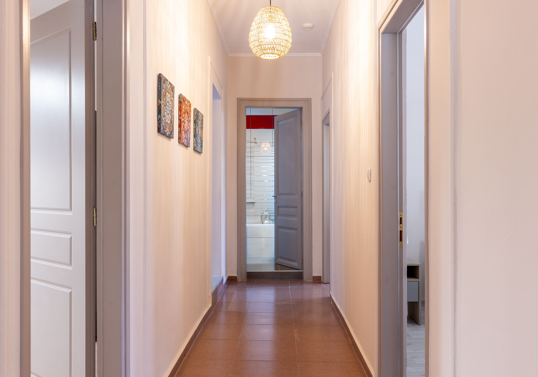 Un corridoio con pareti bianche, pavimento in legno e un lampadario. Ci sono porte su entrambi i lati e una porta in fondo che dà su un bagno.