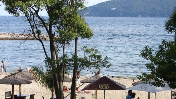 Ein Restaurant direkt am Strand mit Außenbereich, mit Blick auf ein ruhiges Meer und eine hügelige Landschaft im Hintergrund.