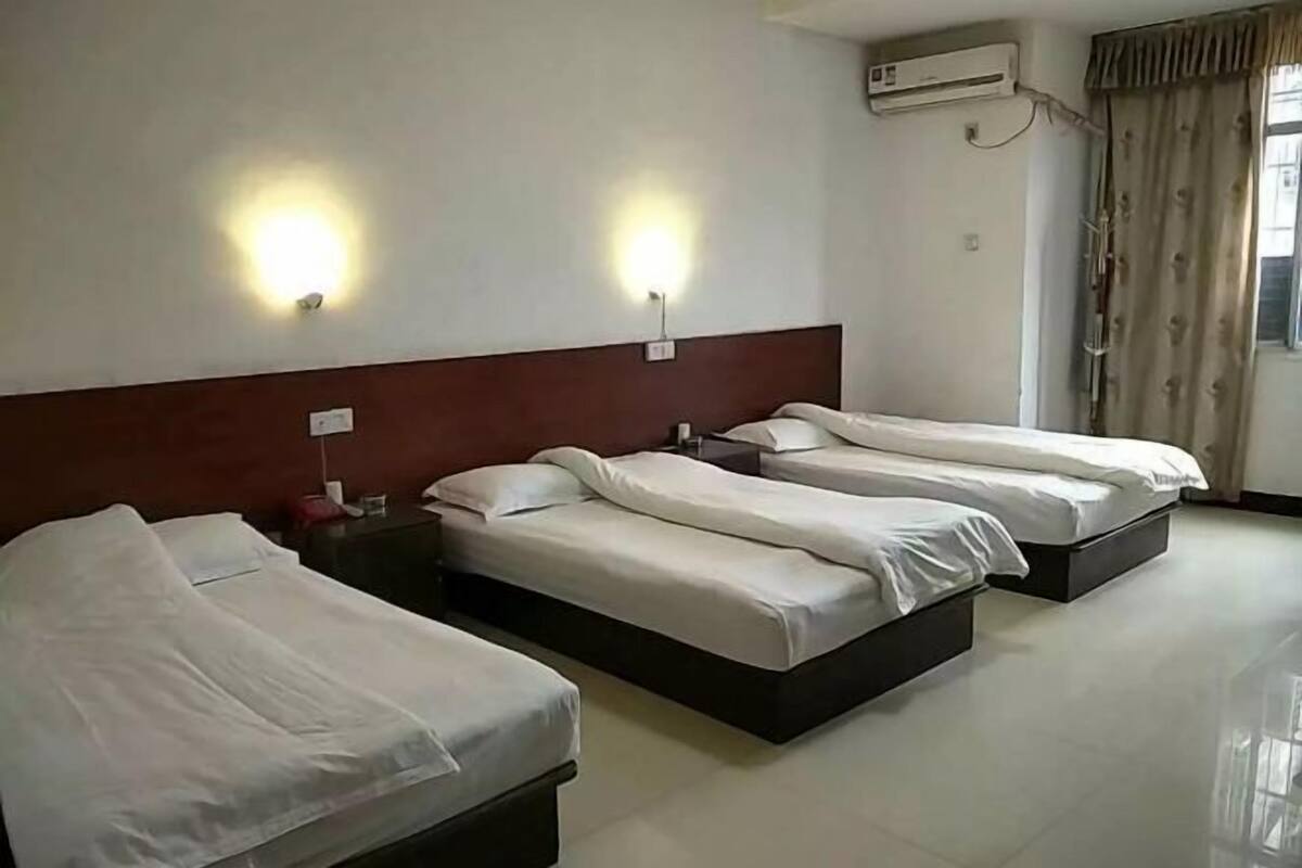 Habitación de hotel con tres camas, una mesita de noche, un equipo de aire acondicionado y una ventana con cortinas.