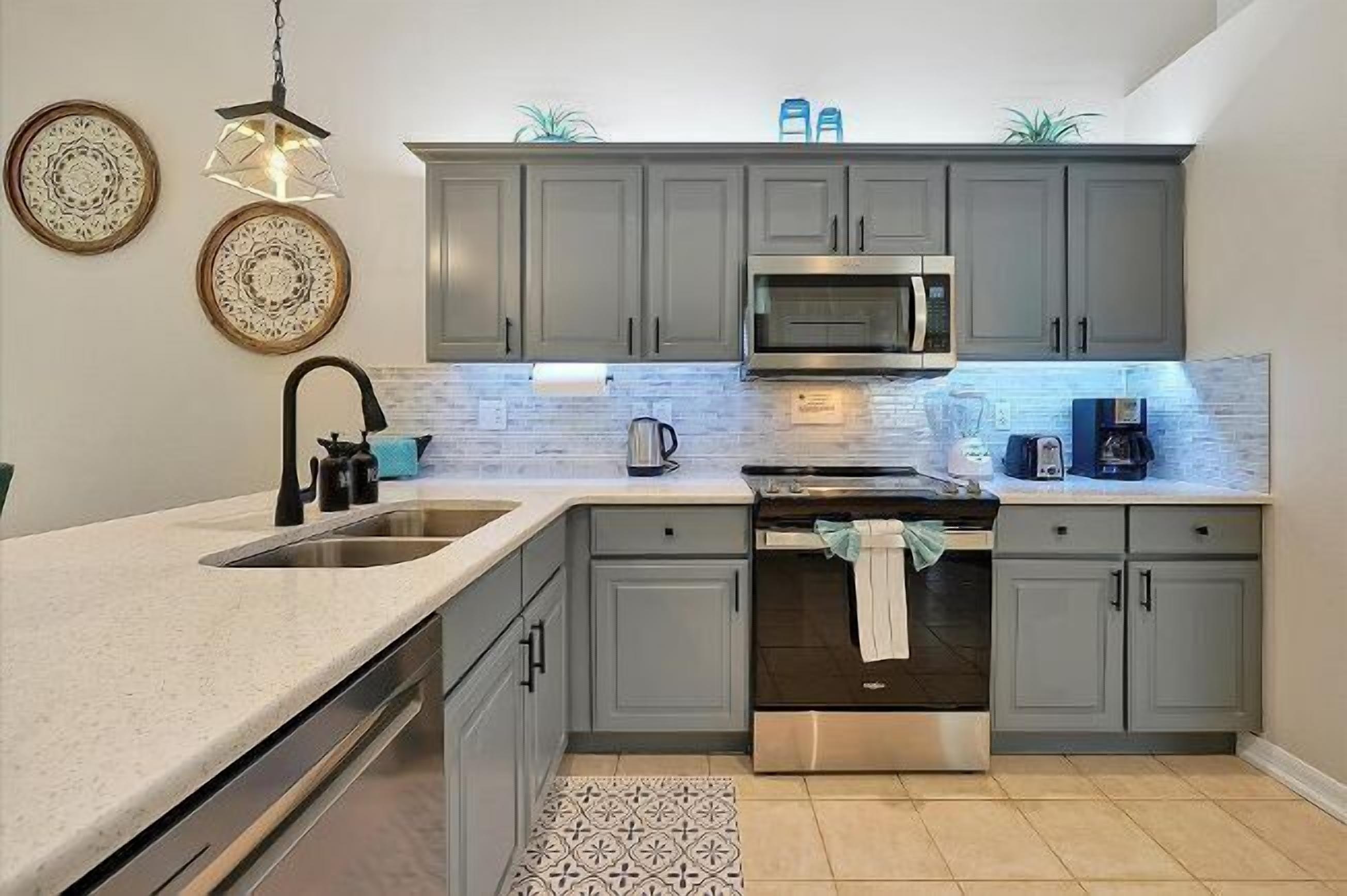 A modern kitchen with grey cabinets, a stainless steel oven, a microwave, and a sink. Decorative plates hang on the wall, and there are two hanging lights.