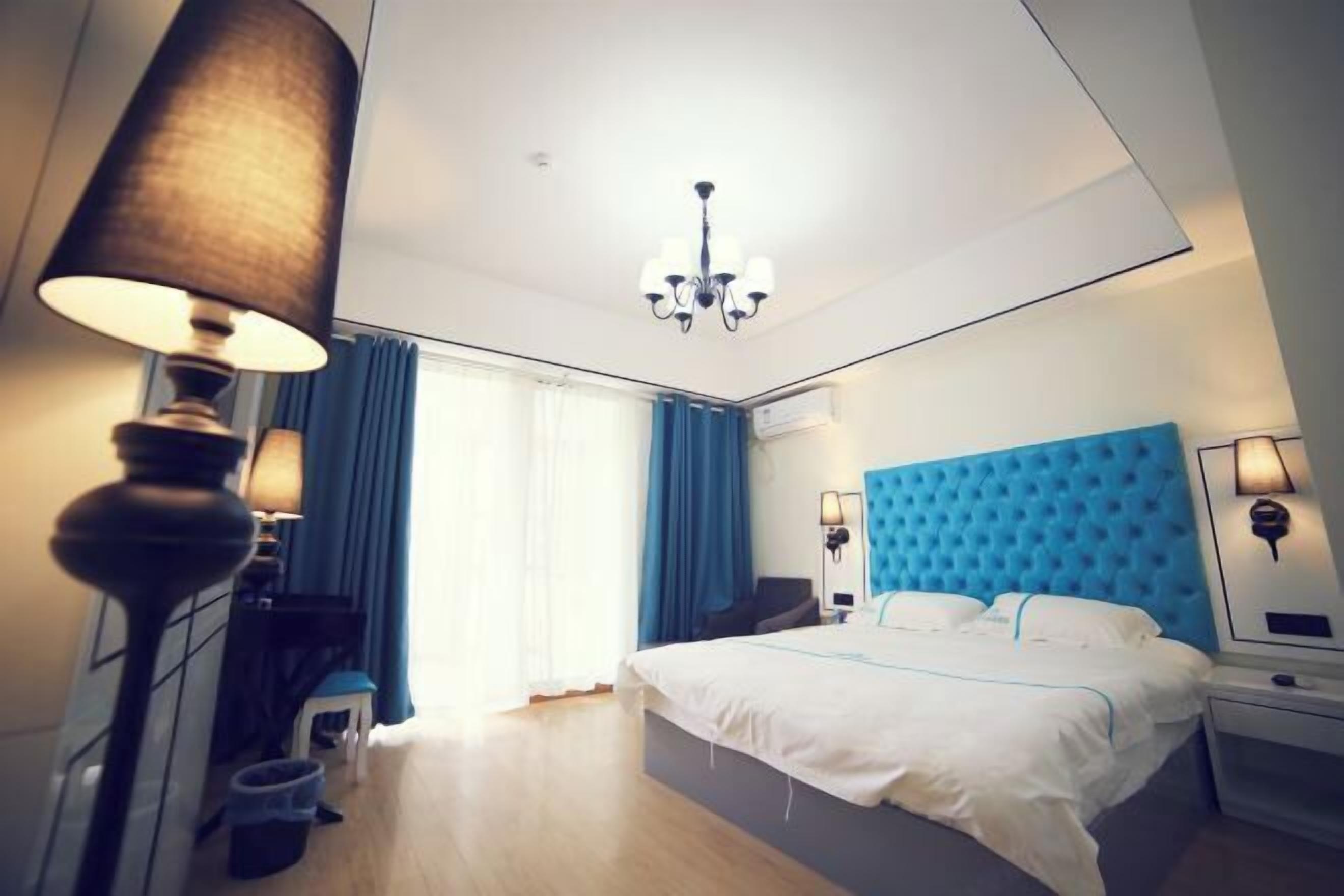 A modern hotel room with a large bed, a chandelier, a blue headboard, and a desk with a lamp.