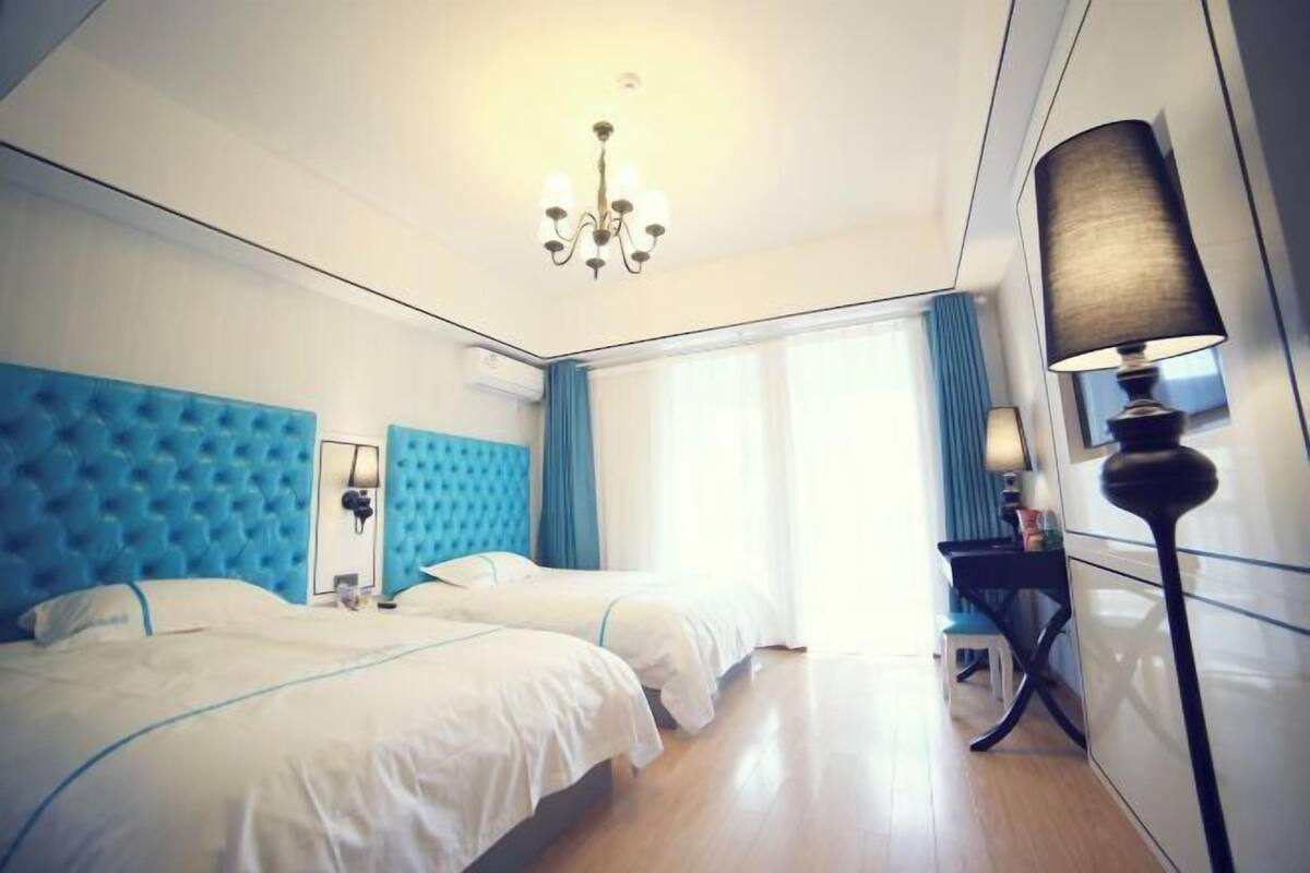 A hotel room with two beds, a chandelier, a desk with a lamp, and a window with curtains.