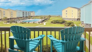 Blick von einem Balkon mit zwei türkisfarbenen Adirondack-Stühlen auf einen Poolbereich mit blauem Himmel und einem fernen Strand.