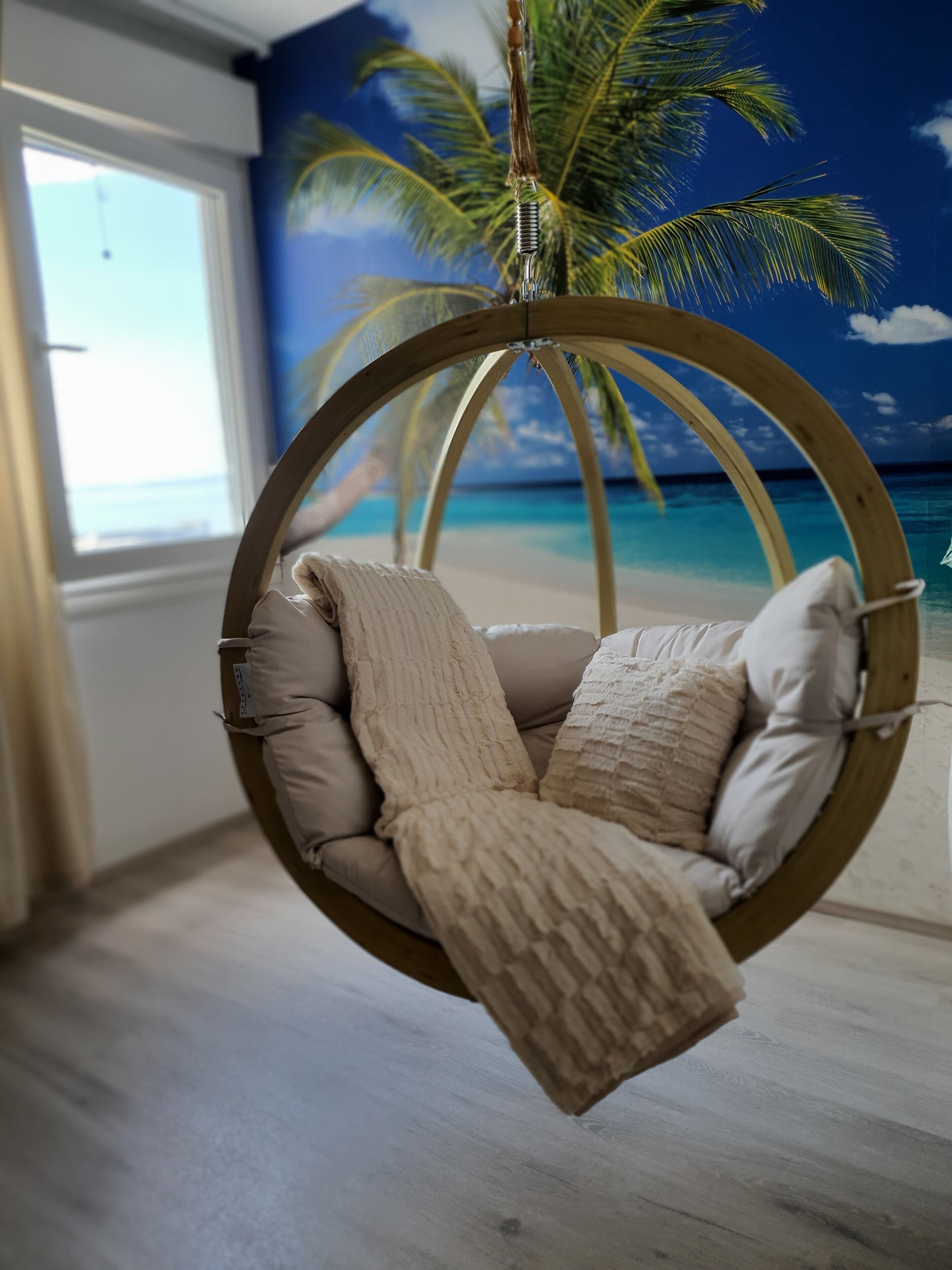 A circular hanging chair with cushions in a room with a beach mural and a window.