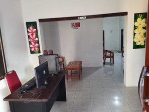 A reception area with a desk, computer, and two speakers. There are two wooden chairs and a small table. The walls have floral paintings.