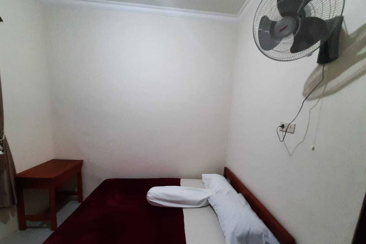 A small, single-bed room with a red bedspread, a white pillow, and a wall-mounted fan.