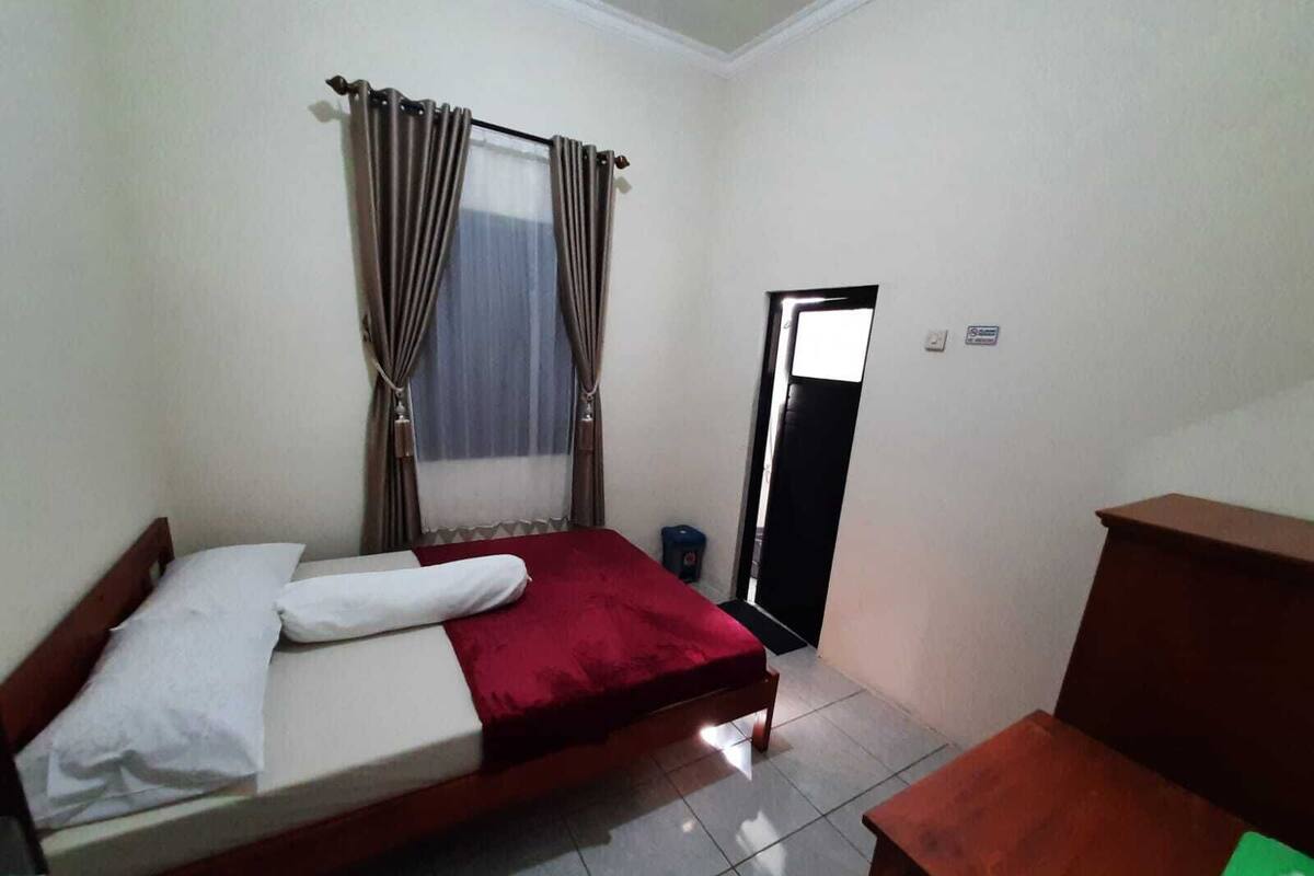 A single-bed room with a wooden headboard, a red bedspread, and a white pillow. There is a small wooden table and a blue trash bin near the door.