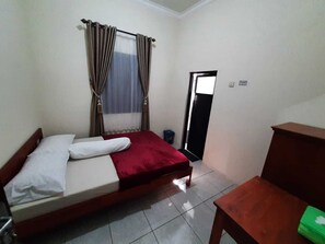A single-bed room with a wooden headboard, a red bedspread, and a white pillow. There is a small wooden table and a blue trash bin near the door.