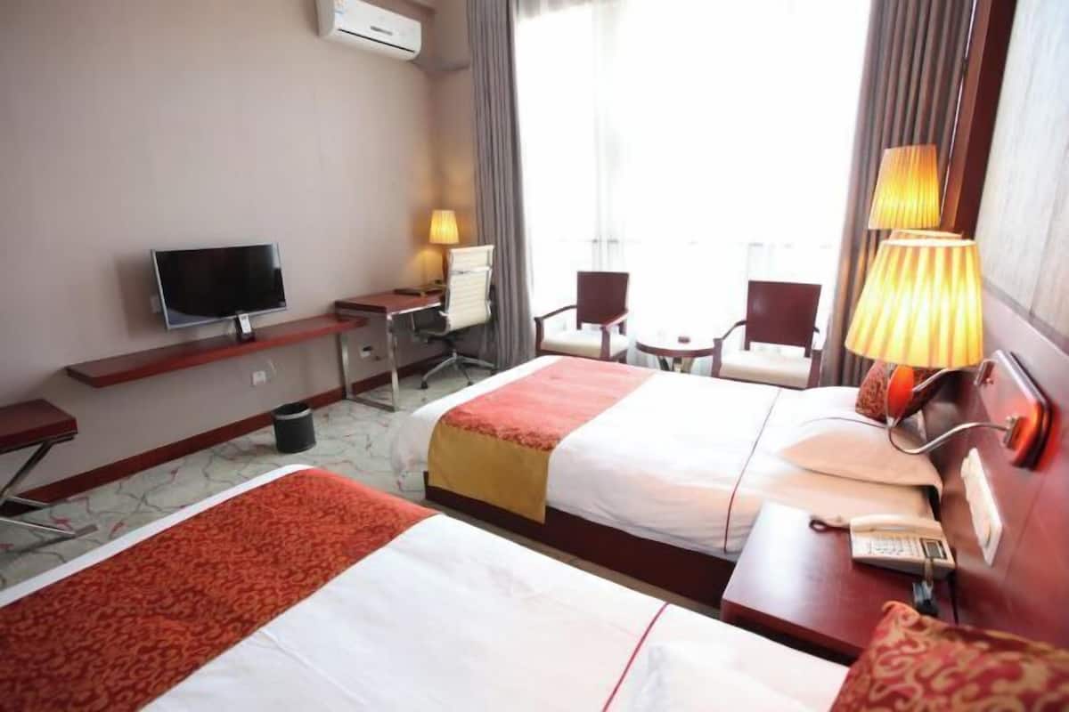 A hotel room with two beds, a desk, a chair, a TV, and a lamp.